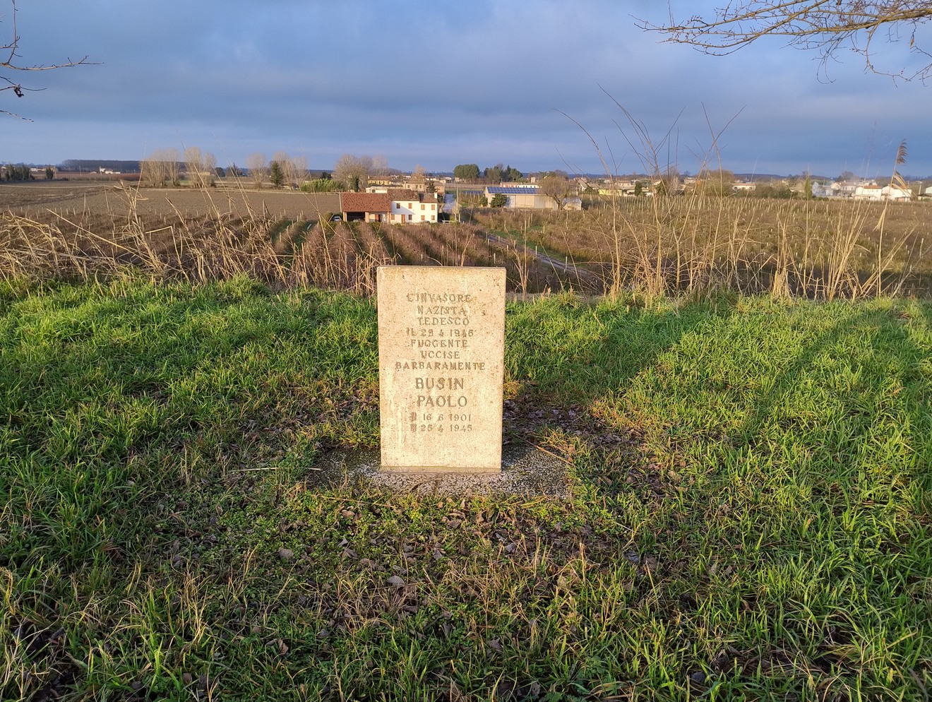 Cippo commemorativo situato a Busin, circondato da un paesaggio rurale e collinare.