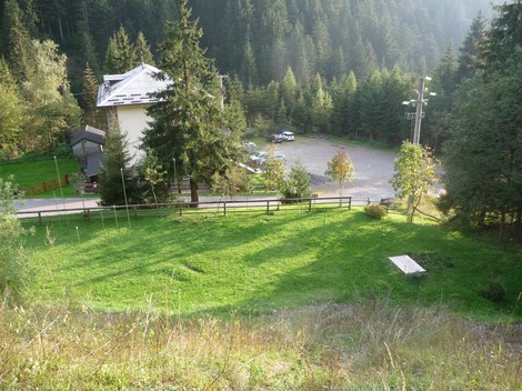 Monumento ai Dodici Caduti immerso nella natura, circondato da alberi e con vista su un parcheggio e un edificio.