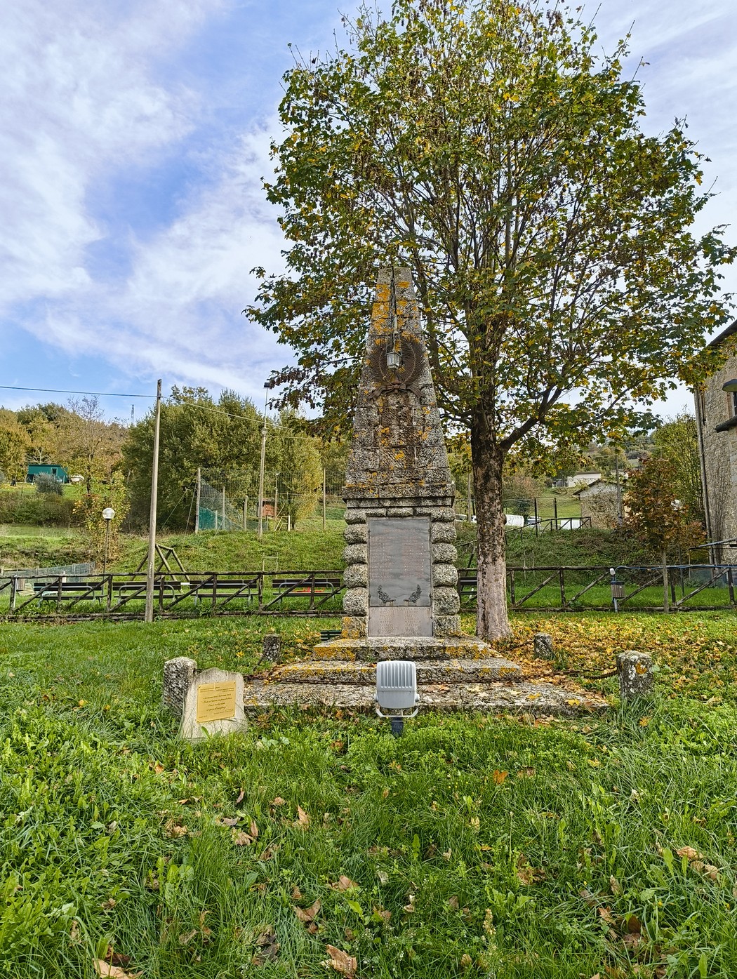 Cippo commemorativo dedicato all'accoglienza degli sfollati, immerso nel verde e circondato da alberi.