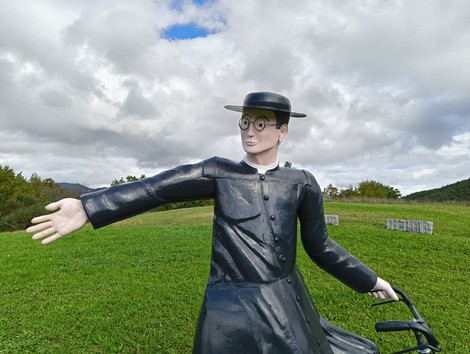 Statua di un angelo in bicicletta, con braccio teso verso il cielo, su un prato verde sotto un cielo nuvoloso.