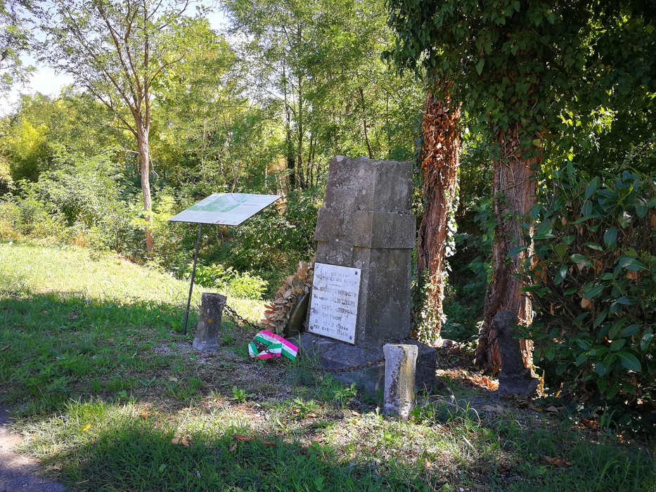 Cippo in pietra con targa e iscrizione, ai bordi di un sentiero nel bosco, con mazzetto tricolore ai piedi.