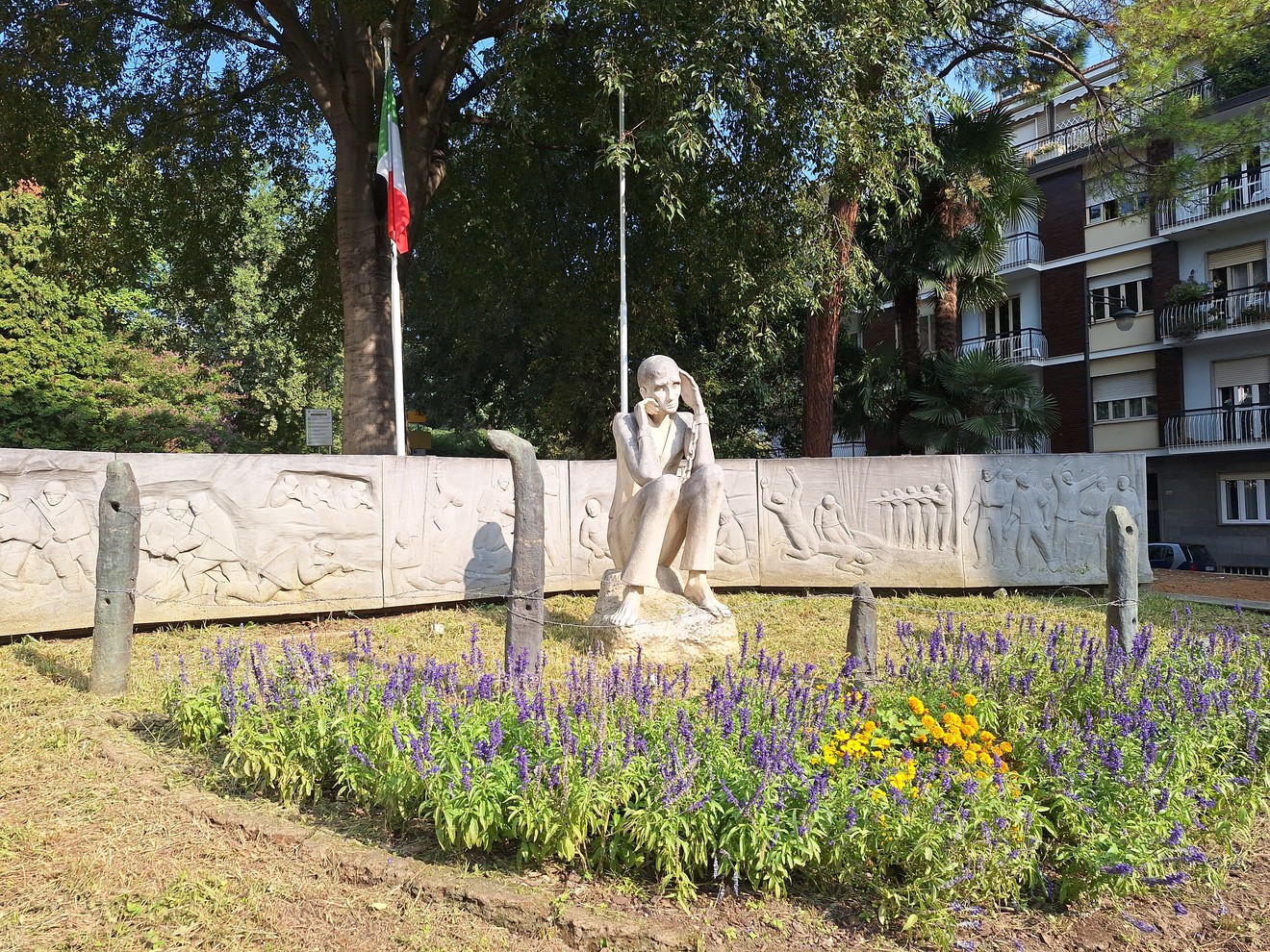 Statua in pietra di una figura seduta davanti a un bassorilievo curvo, tra alberi e aiuole fiorite.