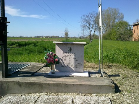 Cippo commemorativo a Beltrami, con fiori e il paesaggio rurale sullo sfondo.