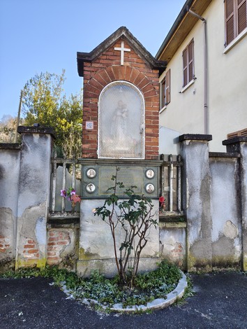 Lapide commemorativa con nicchia decorativa, circondata da un giardino e edifici storici sullo sfondo.