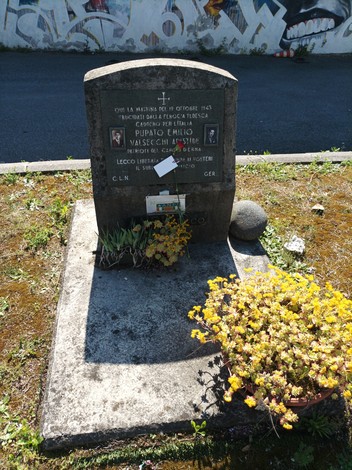 Cippo commemorativo a Pupato e Valsecchi, con fiori e decorazioni, su un fondo di murales.