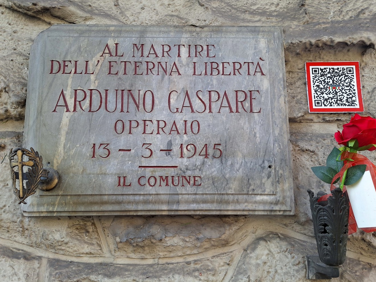 Lapide di marmo con iscrizione su parete in pietra, accanto una rosa rossa in un vaso e un codice QR.