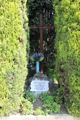 Un monumento commemorativo circondato da arbusti, con una croce e fiori, dedicato ai caduti per la libertà.
