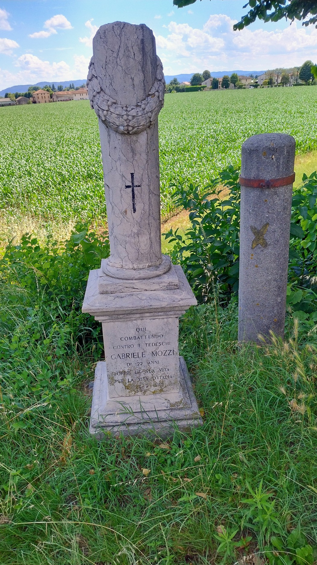 Cippo commemorativo in pietra con decorazioni, immerso in un paesaggio verde di campi e cielo sereno.