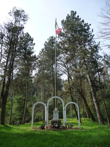 Monumento ai Caduti partigiani circondato da alberi, con una bandiera italiana che sventola in alto.