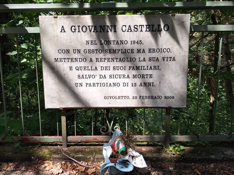 Lapide commemorativa dedicata a Giovanni Castello, con un omaggio floreale in un contesto naturale.