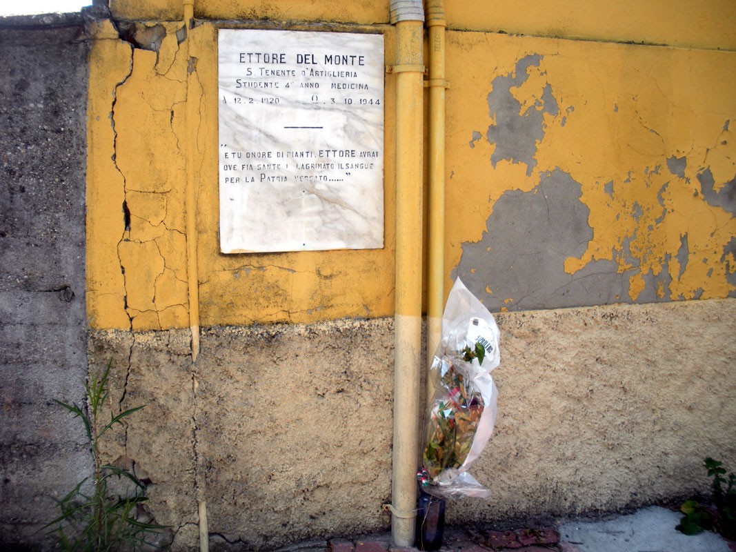 Lapide commemorativa di Ettore Del Monte, con fiori e dedica, su una parete gialla.