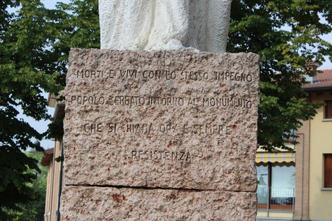 Monumento alla Resistenza, con base in pietra e statua, circondato da alberi e edifici.