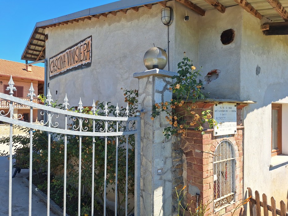 Lapide commemorativa su un muro di una casa, con vegetazione circostante e cancellata in metallo.