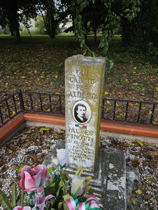 Cippo commemorativo di Finotti, con fiori e decorazioni, immerso nel verde e circondato da una recinzione.