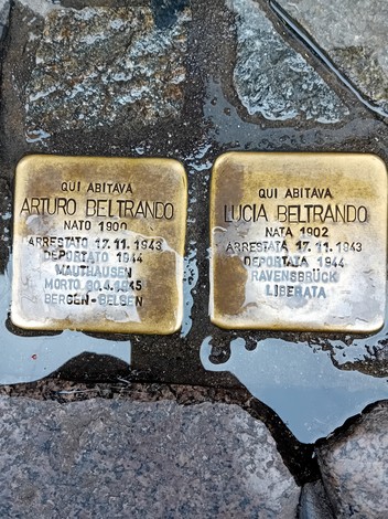 Due pietre d'inciampo in ottone incastonate nel selciato, con iscrizioni incise, bagnate dalla pioggia.