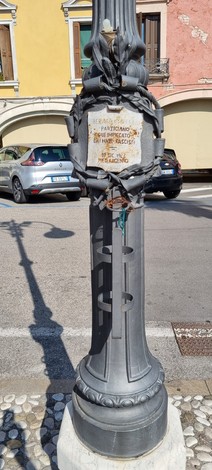 Targa commemorativa su un lampione, decorata con elementi floreali, in una piazza cittadina.