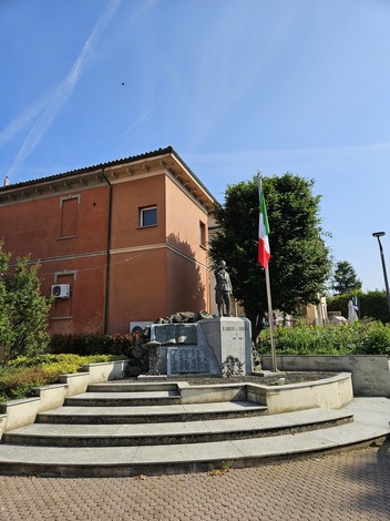 Monumento ai Caduti a San Rocco al Porto, con scalinata, bandiera italiana e sfondo di edifici e verde.