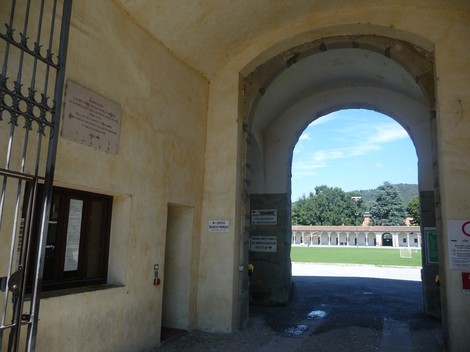 Ingresso con arcata, caratterizzato da una lapide e un cortile visibile sullo sfondo, circondato da alberi.