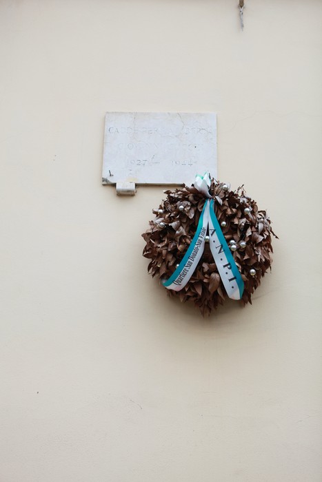 Lapide commemorativa con corona di alloro, appesa a un muro chiaro in un contesto urbano.