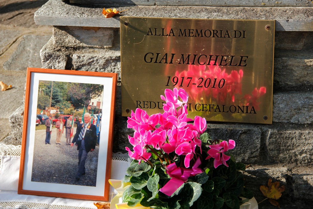 Targa in ottone con iscrizione su muro in pietra, accanto una foto incorniciata e un vaso di ciclamini rosa.