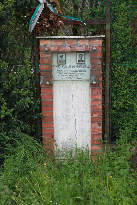 Cippo commemorativo in mattoni dedicato a Simoni Pietro e Arstani Aldo, immerso nella vegetazione.