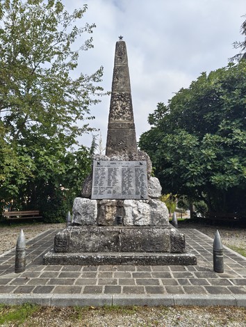 Monumento a obelisco in pietra con lapide incisa, su basamento roccioso in uno spiazzo alberato.