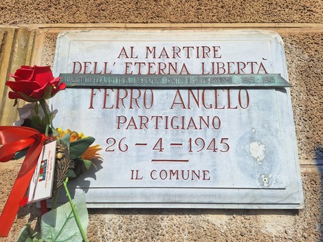 Lapide in marmo su parete esterna, con iscrizione e una rosa rossa deposta accanto.