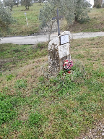 Cippo commemorativo con fiori, situato in un paesaggio collinare e verdeggiante.