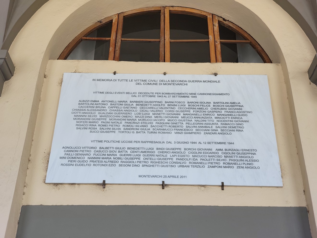 Lapide con iscrizione affissa sopra un portico, sotto una finestra ad arco con persiane in legno.