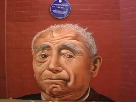 Murale che ritrae un volto umano espressivo, con dettagli sugli occhi e capelli bianchi, su muro rosso.