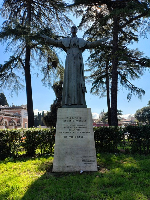 Statua di Eugenio Pacelli con braccia aperte, circondata da alberi e monumenti storici.