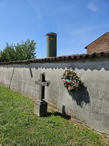 Una croce di legno con una corona di fiori appesa a un muro, sullo sfondo un silos e un cielo azzurro.