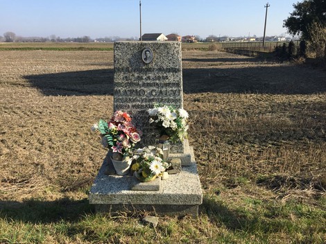 Cippo commemorativo a Calvi, circondato da fiori, in un paesaggio rurale.