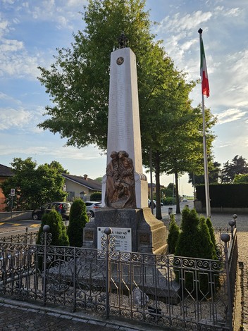Monumento ai Caduti di Pieve Fissiraga, con obelisco e sculture commemorative, circondato da verde e bandiera italiana.