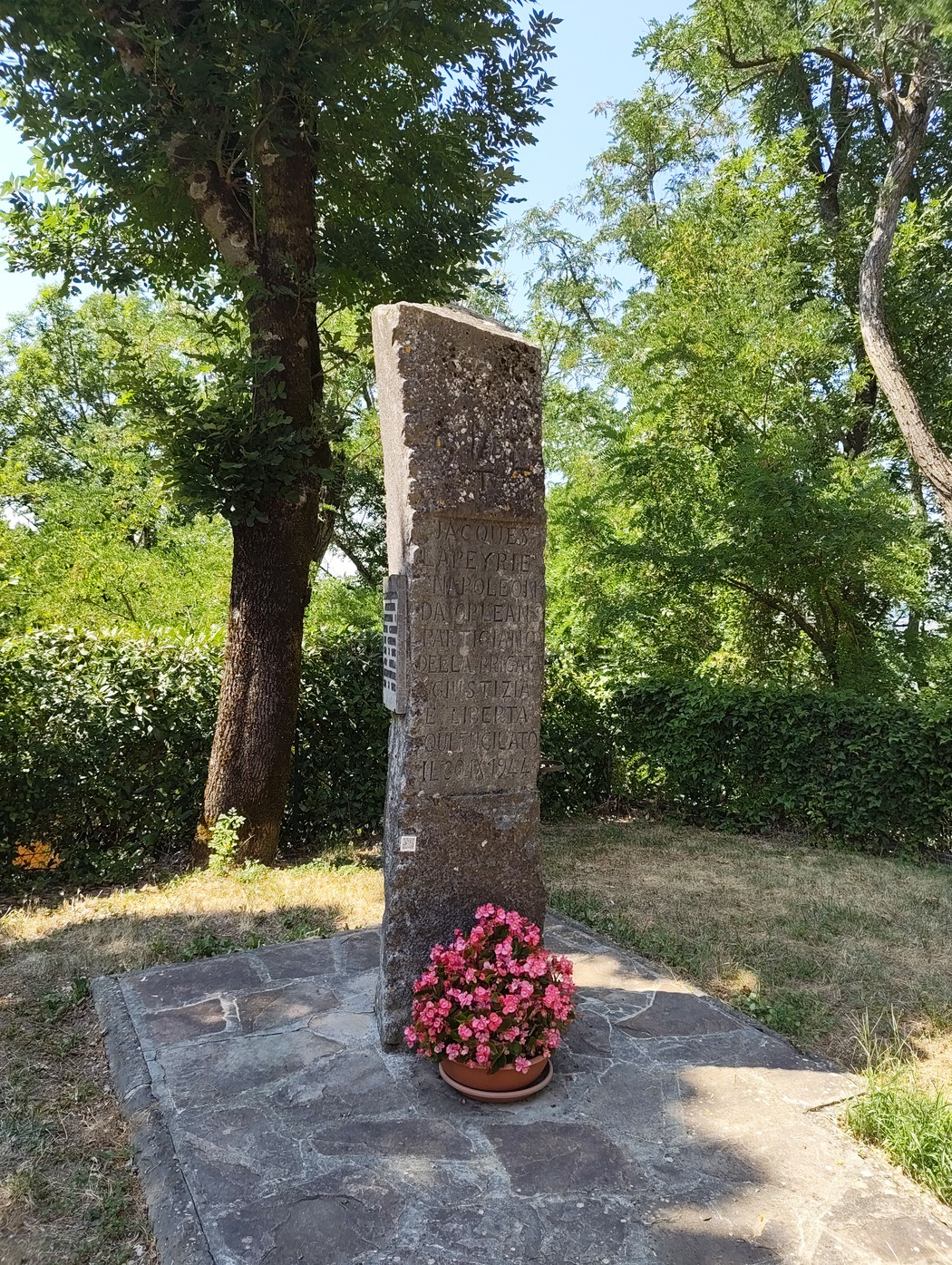 Cippo in pietra con iscrizione, su basamento in un giardino alberato, con vaso di fiori ai piedi.