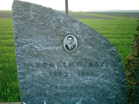 Cippo commemorativo con ritratto ed epigrafe, circondato da un vasto campo verde.