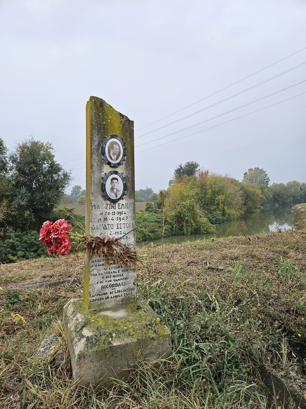 Cippo in pietra con due ritratti fotografici e iscrizione, fiori rossi ai piedi, lungo un canale in campagna.