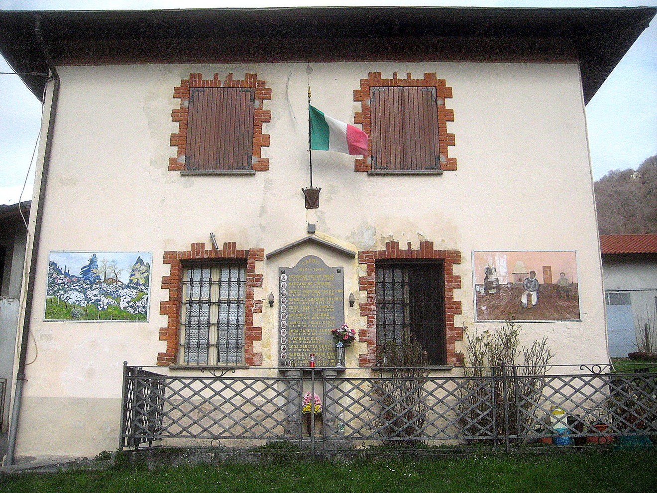 Lapide commemorativa ai caduti di Valle, adornata da fiori e bandiere, su un edificio storicamente significativo.