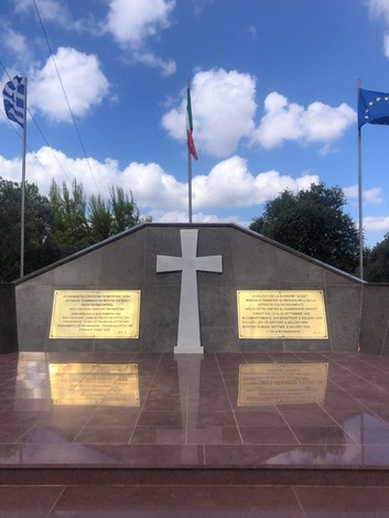 Monumento commemorativo con croce bianca, lastre commemorative e bandiere di Grecia, Italia ed Europa.