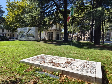 Lastra orizzontale con iscrizione su prato, monumento scultoreo in pietra e alberi sullo sfondo