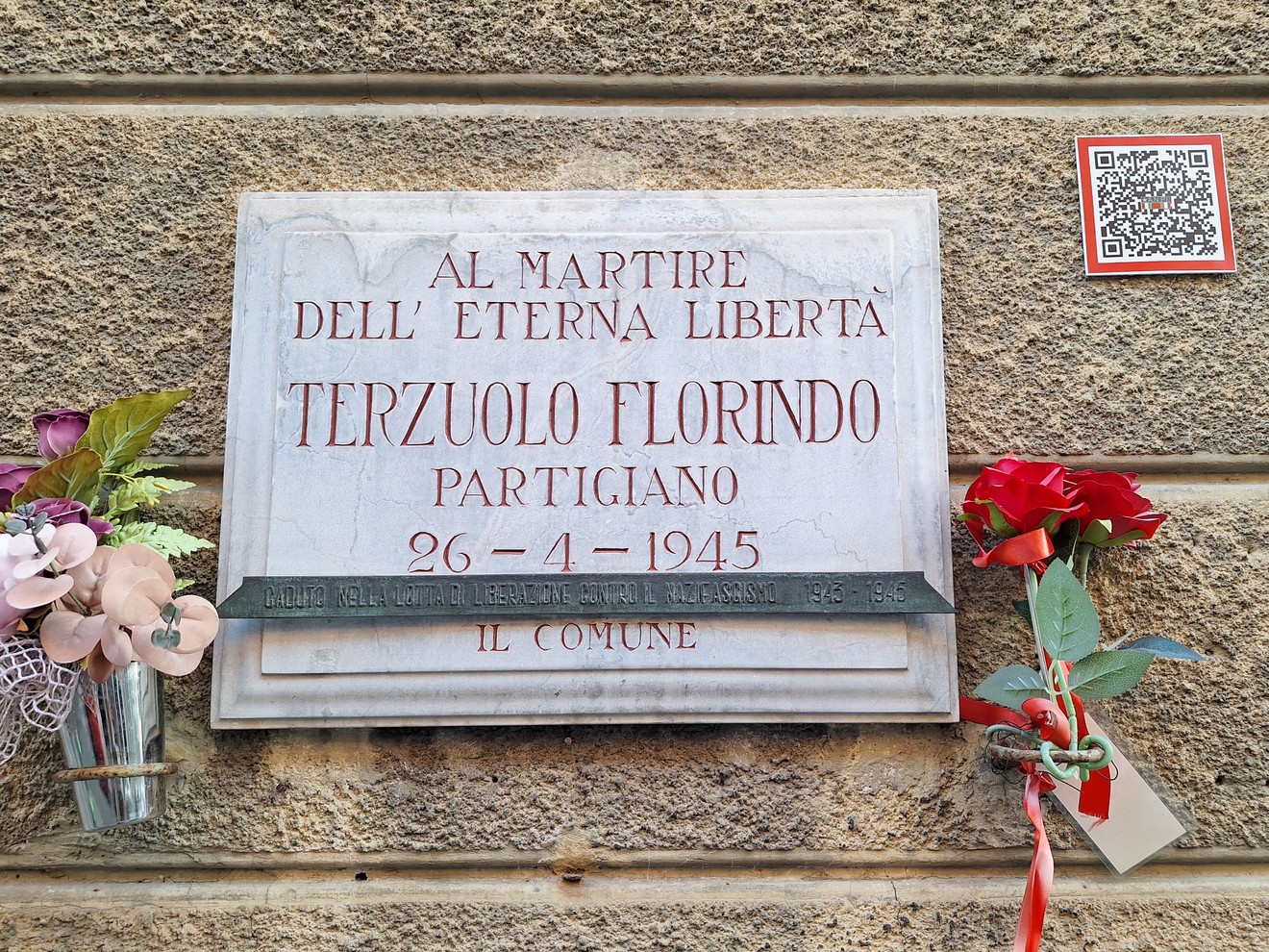 Lapide di marmo con iscrizione su parete, affiancata da fiori freschi e finti.