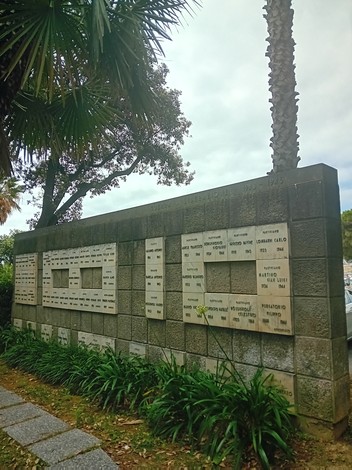 Monumento ai caduti, stone wall with engraved names, surrounded by greenery and palm trees.