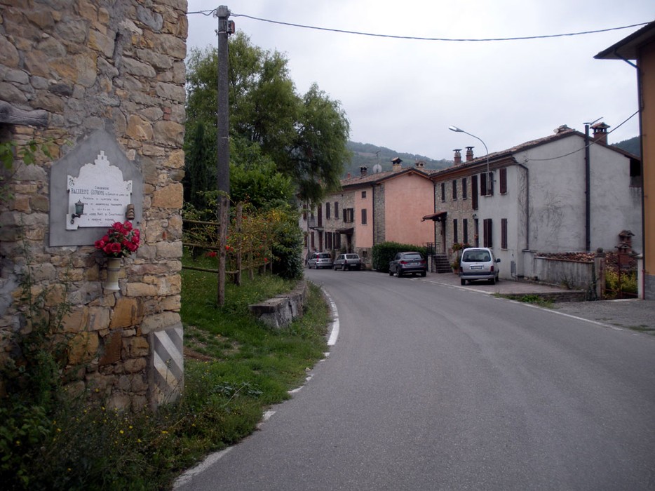 Lapide commemorativa a Ballerini, situata in un tranquillo villaggio. Strada che si snoda tra le abitazioni.