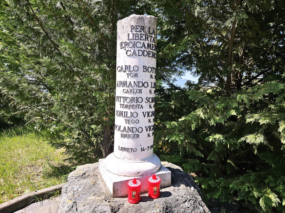 Cippo in pietra a forma di colonna con iscrizione, su basamento roccioso tra alberi di conifere, con due candele rosse ai piedi.