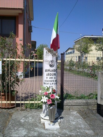 Cippo commemorativo di Armando Arduini con fiori e bandiera italiana, in un contesto urbano.