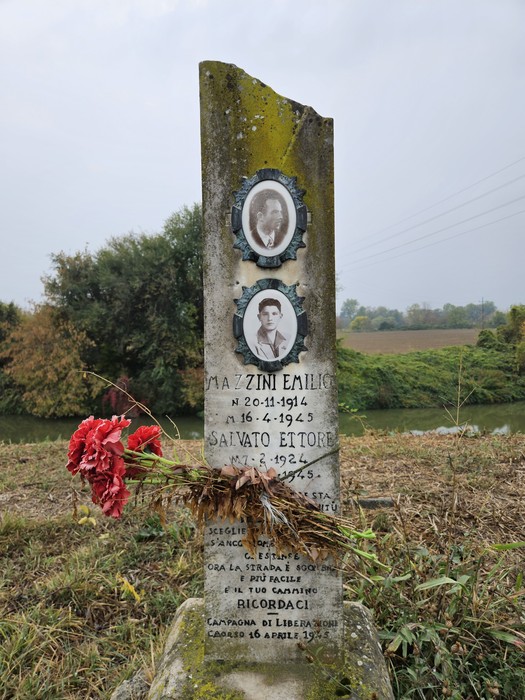 Cippo in pietra con due foto ovali e iscrizione, circondato da campagna, con fiori deposti alla base.