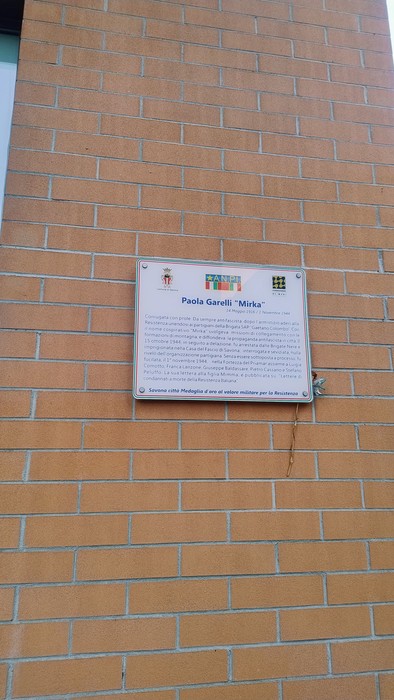 Targa commemorativa su un muro di mattoni, dedicata a Paola Garelli, con informazioni storiche.