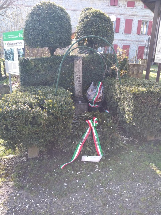 Cippo commemorativo circondato da cespugli ben curati e decorato con nastri tricolori.