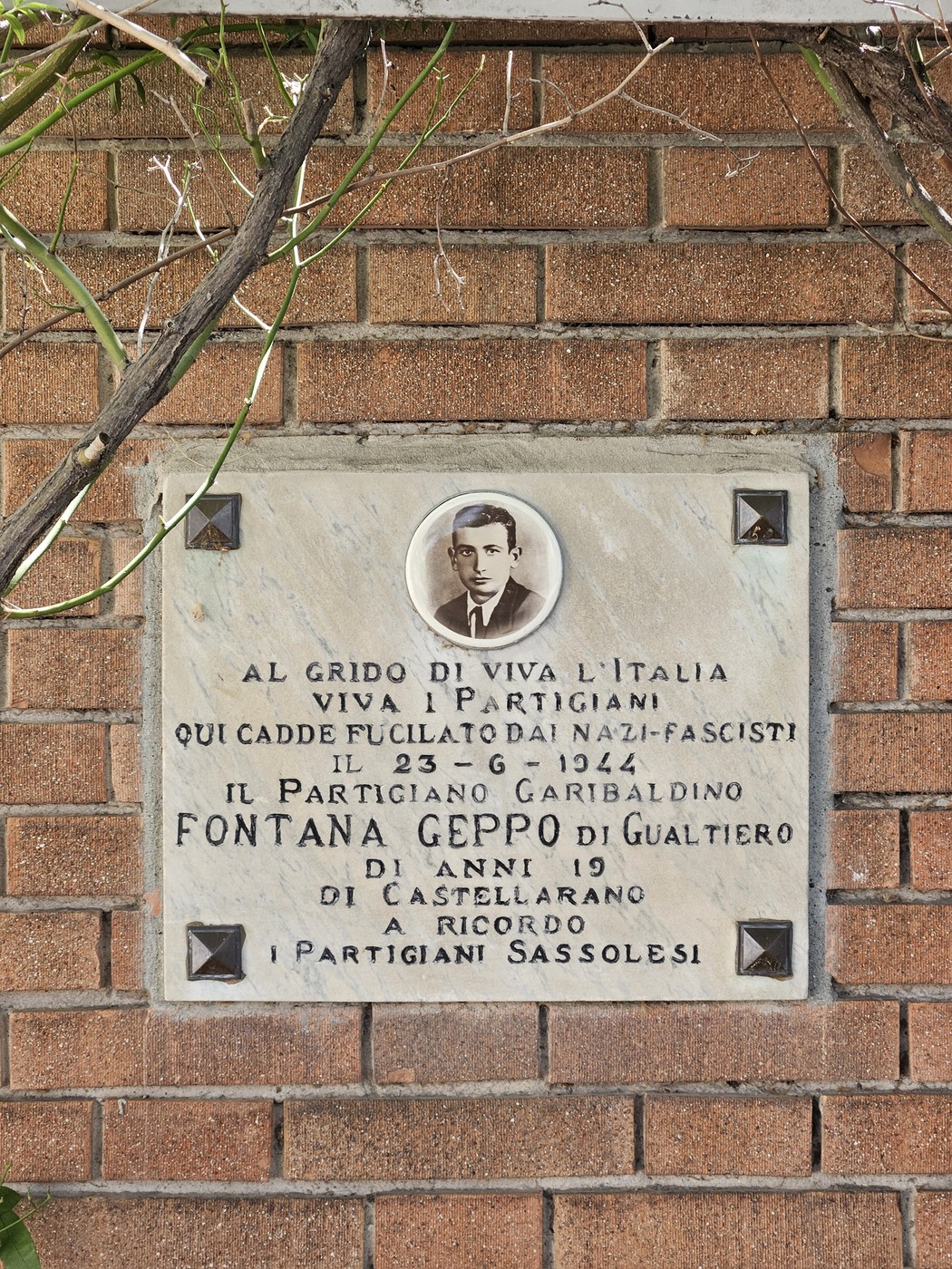 Lapide commemorativa dedicata a Fontana Geppo, partigiano caduto, con decorazioni floreali sullo sfondo.