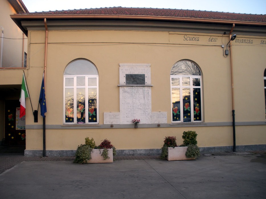 Lapide ai caduti di Robecco Pavese, affissa sulla facciata della scuola, circondata da fiori e bandiere.